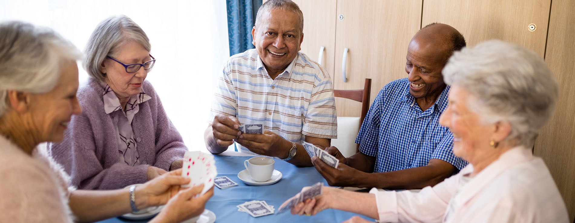 A group of senior residents sitting at a table. The table has a blue cloth covering and a cup of coffee is sitting on it. The senior residents are laughing together while playing cards.